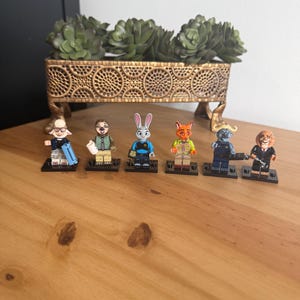 May include: Six Zootopia character minifigures standing on individual black rectangular bases. The figures include a sheep, a sloth, a rabbit police officer, a fox, a buffalo police officer, and a lion, each featuring detailed clothing and accessories.