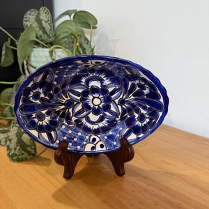 May include: An oval ceramic serving dish in blue and white, decorated with a floral pattern. The dish is displayed on a dark brown wooden stand. The design features stylized flowers and leaves.
