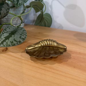 May include: A small, brass-colored ashtray in the shape of a seashell. The ashtray has a ridged interior and a scalloped edge. It sits on a wooden surface, with a potted plant visible in the background.
