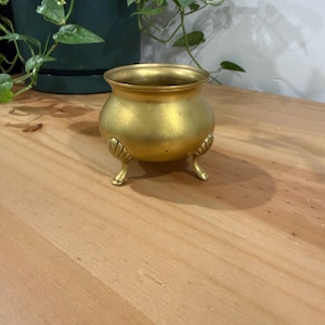Peut inclure: Petit pot en laiton de style antique, avec un corps arrondi et trois petits pieds incurvés. Le pot a une large ouverture et des accents décoratifs en forme de feuilles près de la base. La surface a un aspect légèrement vieilli et texturé.