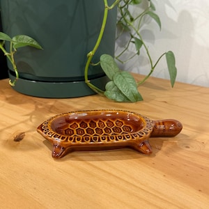 Może przedstawiać: Brązowa, szkliwiona miseczka ceramiczna w kształcie żółwia. Skorupa posiada wypukły, powtarzający się wzór sześciokątów na środku oraz ozdobną obwódkę wokół krawędzi. Naczynie ma cztery małe nóżki i gładkie, błyszczące wykończenie powierzchni.