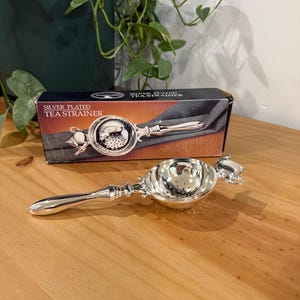 May include: A silver-plated tea strainer with a polished finish, featuring a long handle and a perforated bowl. The strainer is displayed in front of its original rectangular box, which is black and red with the text SILVER PLATED TEA STRAINER.