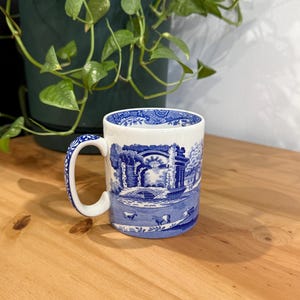 May include: A white ceramic mug with a blue landscape design. The mug features a curved handle and a detailed illustration of a bridge, trees, and animals. The mug is sitting on a wooden surface.