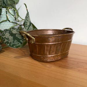 May include: A small, antique copper bucket with two ornate gold-colored handles. The bucket has a ribbed design and a weathered appearance, suggesting age. It sits on a wooden surface, with a leafy green plant visible in the background.