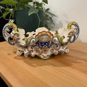 May include: A white ceramic centerpiece bowl featuring ornate, colorful hand-painted scrollwork in shades of blue, yellow, orange, and green. The bowl has two curved handles with sculpted faces and a scalloped rim, supported by small feet.