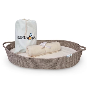 May include: A brown oval-shaped woven basket with a white cotton liner and a white rolled towel with a white ribbon. The basket has two handles and a white drawstring bag with the text "nova bee" in black lettering and a bee logo.