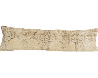 May include: A long, rectangular lumbar pillow featuring a beige fabric base with a faded, intricate dark brown floral and geometric pattern across the entire surface. The pillow has a textured appearance and a soft, slightly uneven rectangular shape.