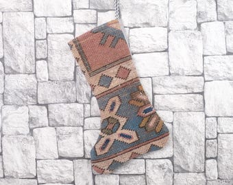 May include: A patterned Christmas stocking with a woven design. The stocking displays a mix of colours, including red, blue, and beige, with geometric and floral motifs. It hangs against a textured, light-coloured stone wall.