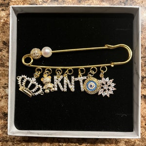 May include: Gold safety pin with charms, including a crown, bear, letters spelling "KNT", an evil eye, and a snowflake. The charms are adorned with clear and blue crystals.