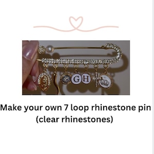 May include: A gold-colored safety pin with clear rhinestones and dangling charms. The pin has a pearl accent and holds seven charms, including a cross, a Virgin Mary pendant, and charms with the letters "GH". The text below reads "Make your own 7 loop rhinestone pin (clear rhinestones)".
