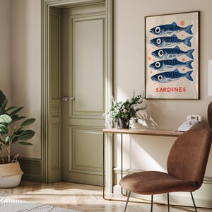 Może przedstawiać: Plakat w stylu vintage z białym tłem i czerwonymi k&oacute;łkami. Pięć niebieskich sardynek ułożonych pionowo, z napisem "SARDINES" nadrukowanym na czerwono poniżej. Plakat jest oprawiony i wisi na ścianie nad drewnianym stolikiem konsolowym.