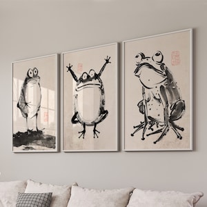 May include: Three black and white ink drawings of frogs in different poses framed in white frames. The frames are hanging on a light gray wall.