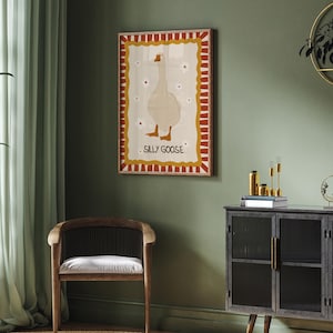 May include: A framed illustration of a white goose with red, yellow, and brown accents. The illustration is titled "Sally Goose". The frame is hanging on a green wall with a dark wood cabinet and a chair in the foreground.