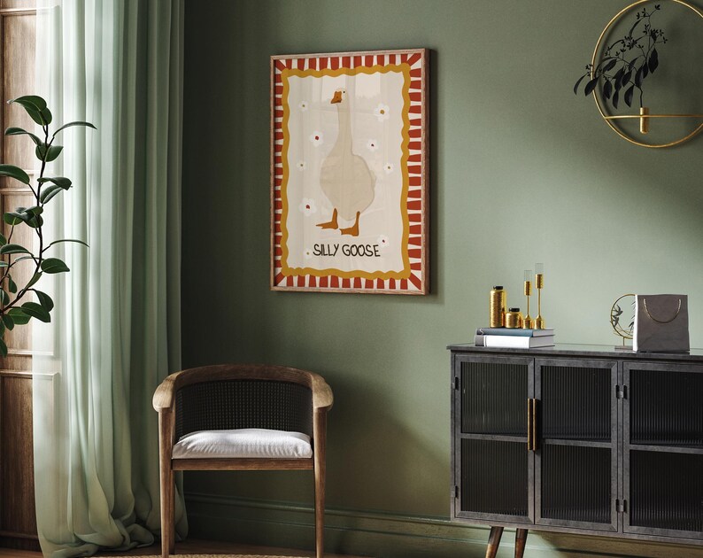 May include: A framed illustration of a white goose with red, yellow, and brown accents. The illustration is titled "Sally Goose". The frame is hanging on a green wall with a dark wood cabinet and a chair in the foreground.