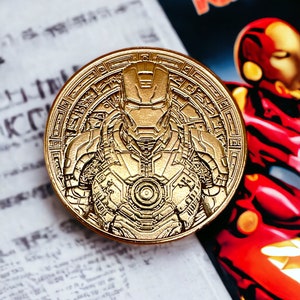 Może przedstawiać: Złota moneta z szczegółowym reliefem zbroi Iron Mana. Moneta ma skomplikowane wzory obramowania i jest umieszczona na tle stron komiksów oraz czerwono-złotej ilustracji.