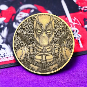 May include: A gold-colored, circular coin featuring a detailed relief of the Deadpool character. The coin has a textured border and the character is depicted with his mask and two swords crossed behind him. The coin rests on a purple surface.
