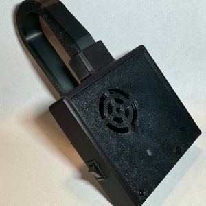 May include: A black, rectangular electronic device with a handle. The device has a circular vent, a small switch, and two screws. The handle is a curved, rectangular shape. The device is likely a small electronic component or tool.