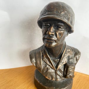 May include: A bronze bust of a man wearing a military helmet and uniform. The sculpture has a detailed face and is mounted on a circular base. The bust is a dark bronze color and sits on a wooden surface.