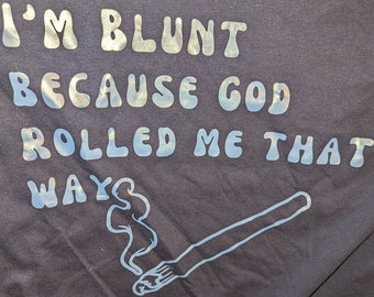 May include: A dark blue t-shirt with the text "I'M BLUNT BECAUSE GOD ROLLED ME THAT WAYS" in a light blue, bubble-like font. A stylized image of a lit joint with smoke is below the text.