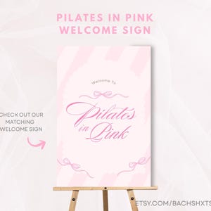 May include: A pink welcome sign on a wooden easel. The sign reads "Pilates in Pink" in elegant script, with decorative bows. The top of the sign says "Pilates in Pink Welcome Sign". The sign also says "Check out our matching welcome sign".