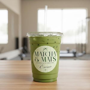 Puede incluir: Un vaso de plástico transparente lleno de bebida de matcha verde y cubitos de hielo, coronado con una pajita transparente. Una pegatina circular blanca en la taza dice "Matcha & Mats" con el texto "A Birthday Celebrating Emma". La taza está sobre una superficie de madera.
