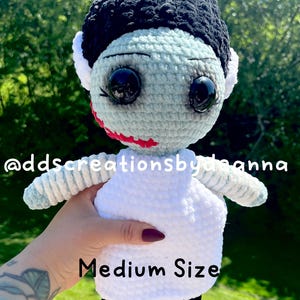 May include: A handmade crochet doll resembling the Bride of Frankenstein. The doll has a light blue face, black hair, and a white dress. The doll's eyes are large and black, and the lips are red. The text "Medium Size" is visible.