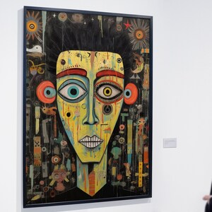 May include: A colorful abstract painting of a face with large eyes, a red mouth, and a yellow background. The painting is surrounded by a black frame and is hanging on a white wall.