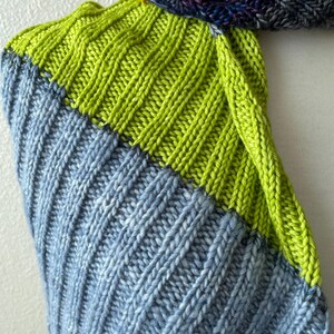 May include: A close-up of a hand-knitted scarf with a ribbed pattern. The scarf features sections of bright green, light blue, and a dark blue and purple section. The knitted texture is clearly visible.