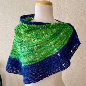 May include: A handmade shawl in shades of green and blue, displayed on a mannequin. The shawl features a gradient design, transitioning from green to blue. The knitted fabric has a textured appearance with openwork details, and is designed to be worn around the neck and shoulders.