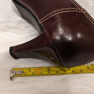 Pu&ograve; includere: Primo piano di una scarpa con tacco in pelle marrone con cuciture bianche. Un metro a nastro giallo &egrave; posizionato sotto il tacco, mostrando una misura di 6,35 cm.