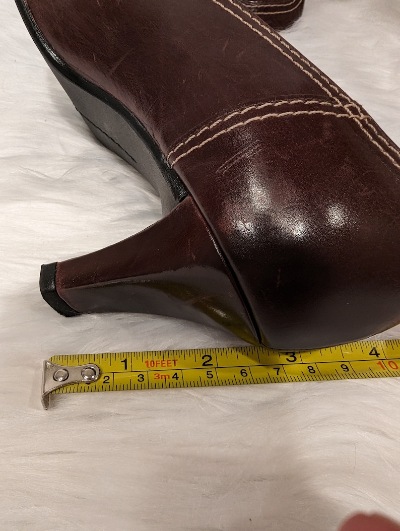 Pu&ograve; includere: Primo piano di una scarpa con tacco in pelle marrone con cuciture bianche. Un metro a nastro giallo &egrave; posizionato sotto il tacco, mostrando una misura di 6,35 cm.