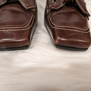 Pu&ograve; includere: Un paio di scarpe in pelle marrone con punta quadrata e un dettaglio di cucitura bianca. Le scarpe hanno un dettaglio a fiocco sulla parte superiore.