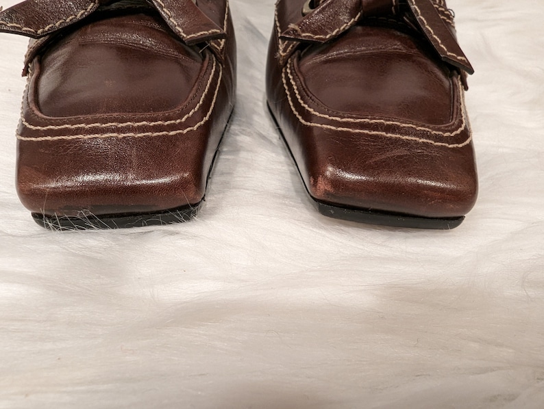 Pu&ograve; includere: Un paio di scarpe in pelle marrone con punta quadrata e un dettaglio di cucitura bianca. Le scarpe hanno un dettaglio a fiocco sulla parte superiore.