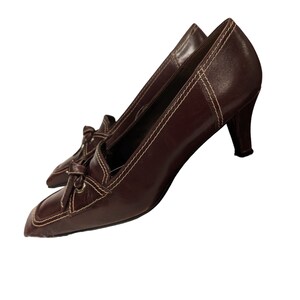 Pu&ograve; includere: Un paio di scarpe con tacco in pelle marrone con punta quadrata e un nodo decorativo sul davanti. Le scarpe hanno un dettaglio di cucitura bianca attorno ai bordi.