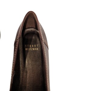 Pu&ograve; includere: Primo piano dell'interno di una scarpa in pelle marrone con il nome del marchio "STUART WEITZMAN" cucito in lettere dorate.