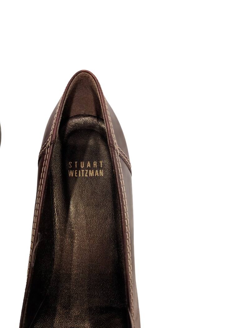 Pu&ograve; includere: Primo piano dell'interno di una scarpa in pelle marrone con il nome del marchio "STUART WEITZMAN" cucito in lettere dorate.