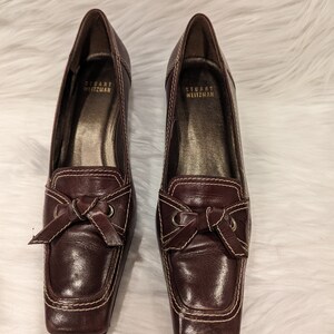 Pu&ograve; includere: Un paio di scarpe con tacco in pelle marrone con un dettaglio a fiocco e cuciture bianche. Le scarpe sono di Stuart Weitzman.