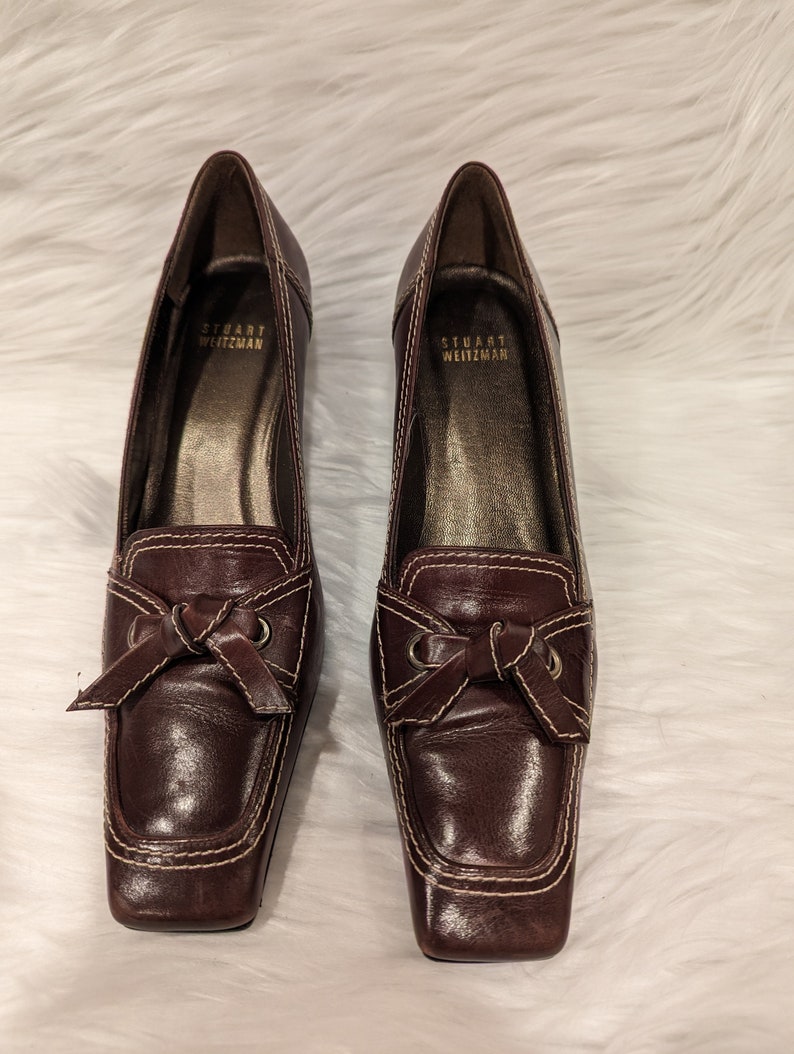 Pu&ograve; includere: Un paio di scarpe con tacco in pelle marrone con un dettaglio a fiocco e cuciture bianche. Le scarpe sono di Stuart Weitzman.