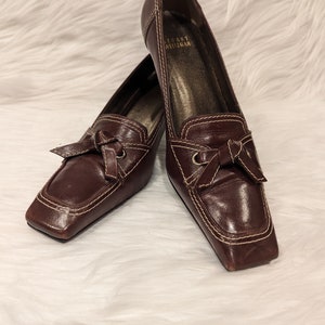 Pu&ograve; includere: Un paio di scarpe con tacco in pelle marrone con punta quadrata e dettaglio a fiocco. Le scarpe sono del marchio "Tory Burch".
