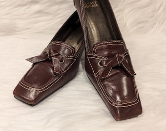 Puede incluir: Un par de zapatos de tacón de cuero marrón con punta cuadrada y detalle de lazo. Los zapatos son de la marca "Tory Burch".