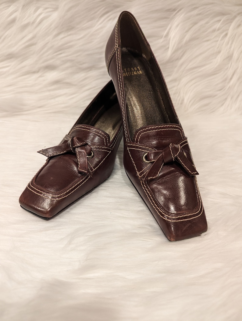 Pu&ograve; includere: Un paio di scarpe con tacco in pelle marrone con punta quadrata e dettaglio a fiocco. Le scarpe sono del marchio "Tory Burch".