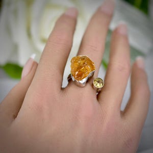 Puede incluir: Un anillo de plata con una gema de citrino en bruto de color naranja dorado engastada en un bisel de metal texturizado, junto a una gema pequeña, redonda y facetada de color amarillo pálido en una banda de plata sencilla.