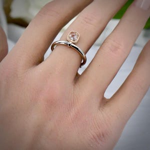 May include: A ring featuring a square-shaped, faceted clear stone set in a rose gold bezel with a twisted rope texture. The band is made of polished silver-toned metal, creating a two-tone design that sits on the finger with a delicate appearance.