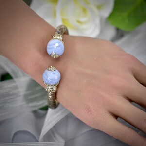 Puede incluir: Una pulsera tipo brazalete con una banda metálica texturizada y remates dorados ornamentados en los extremos. Cada remate está coronado por una cuenta de ágata azul claro, redonda y lisa, con vetas blancas naturales visibles.