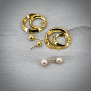 Può includere: Una collezione di gioielli che include due grandi orecchini a cerchio color oro, due piccoli orecchini a lobo color oro e una spilla color oro con due perle sferiche rosa pallido alle estremità, il tutto disposto su una superficie testurizzata.