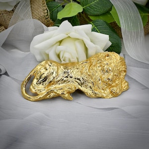 Pode incluir: Uma estatueta de leão em metal dourado representada em posição reclinada com textura de pelo detalhada e uma juba proeminente. O leão está posicionado horizontalmente, exibindo uma superfície reflexiva e detalhes esculturais intrincados.