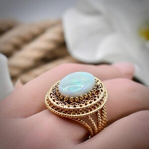 May include: A gold ring featuring a large, oval-shaped cabochon opal stone with iridescent flashes of color. The setting is crafted with intricate, filigree-style metalwork and a twisted rope border, supported by a multi-strand twisted gold band.