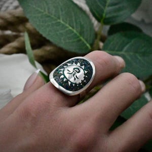 May include: A silver ring featuring a large, rounded, shield-shaped face with a polished silver sun emblem centered against a dark, textured, multi-colored inlay of crushed stone. The band is smooth and tapers toward the back of the finger.
