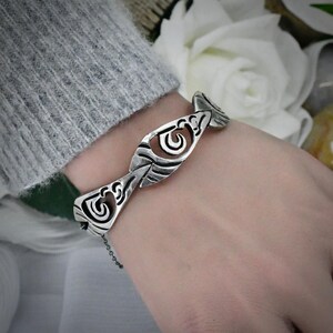 May include: A silver-toned metal bracelet featuring a series of interconnected, stylized geometric links with cut-out spiral patterns. The bracelet has a polished finish with darkened recessed areas to highlight the intricate, repeating design.