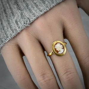 Puede incluir: Un anillo de oro con una pieza central de camafeo de forma ovalada. El camafeo muestra un relieve de perfil blanco de una persona sobre un fondo marrón, engastado en un bisel de oro pulido. La banda es fina y metálica, rodeando el dedo.